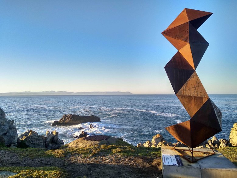 Ucu (2019), a rusted mild steel sculpture inspired by the necklace worn by migrant labourers, using the geometry of triangles to create a balancing act on a cliff in Hermanus.
