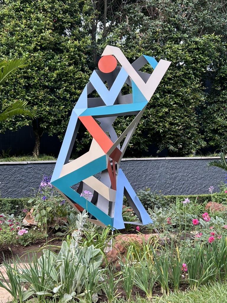 A colourful geometric steel sculpture made of interlocking triangular shapes stands in a landscaped garden with flowers and greenery beside a pool and a tall green wall.